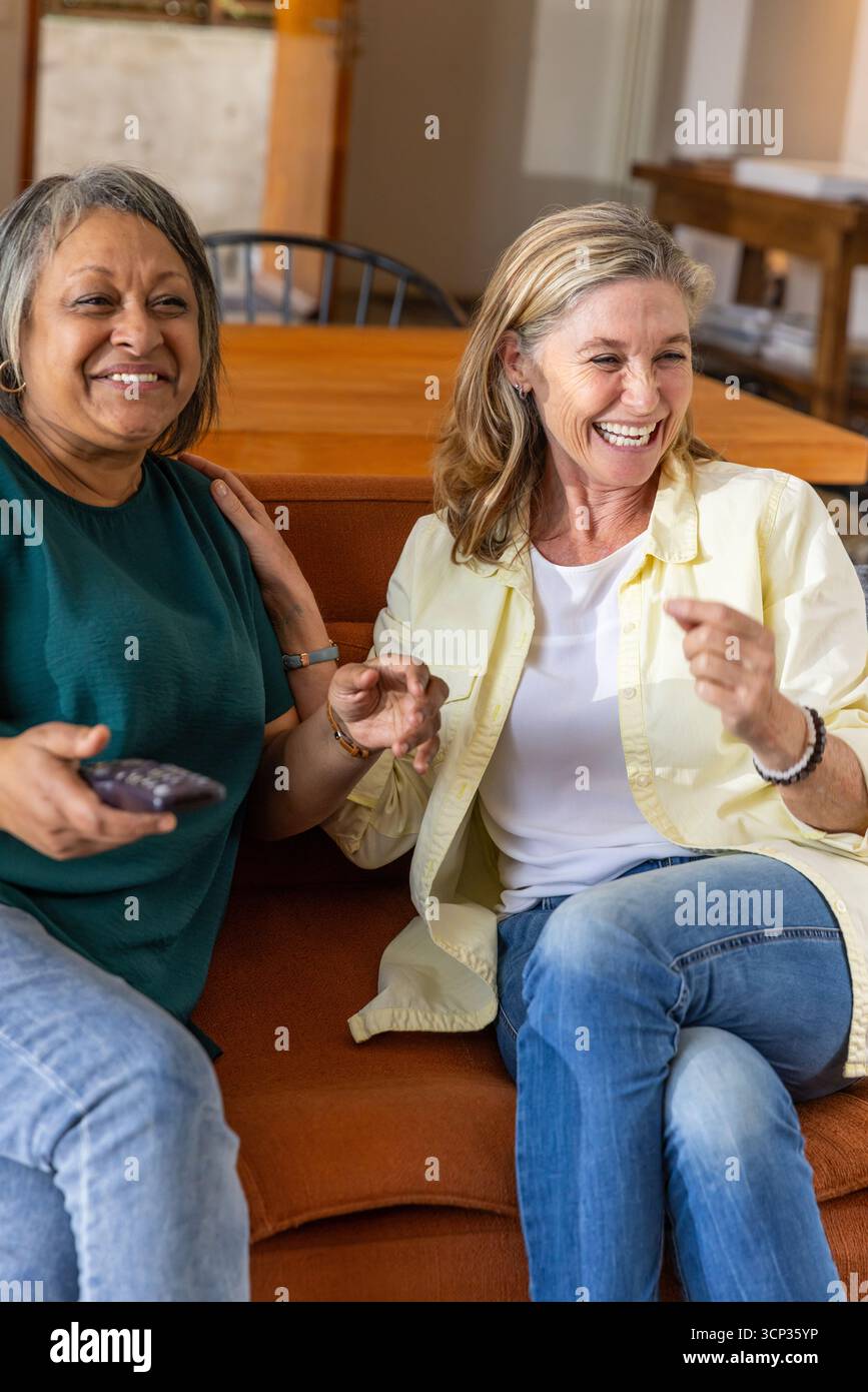 Amici donne anziane che chattano animatamente sul divano del soggiorno con telecomando tv Foto Stock