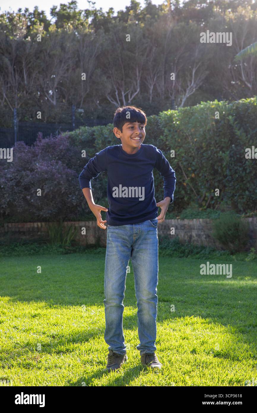 Ragazzo indiano in piedi su un prato erboso nel giardino sul retro con pareti in mattoni, siepe densa Foto Stock