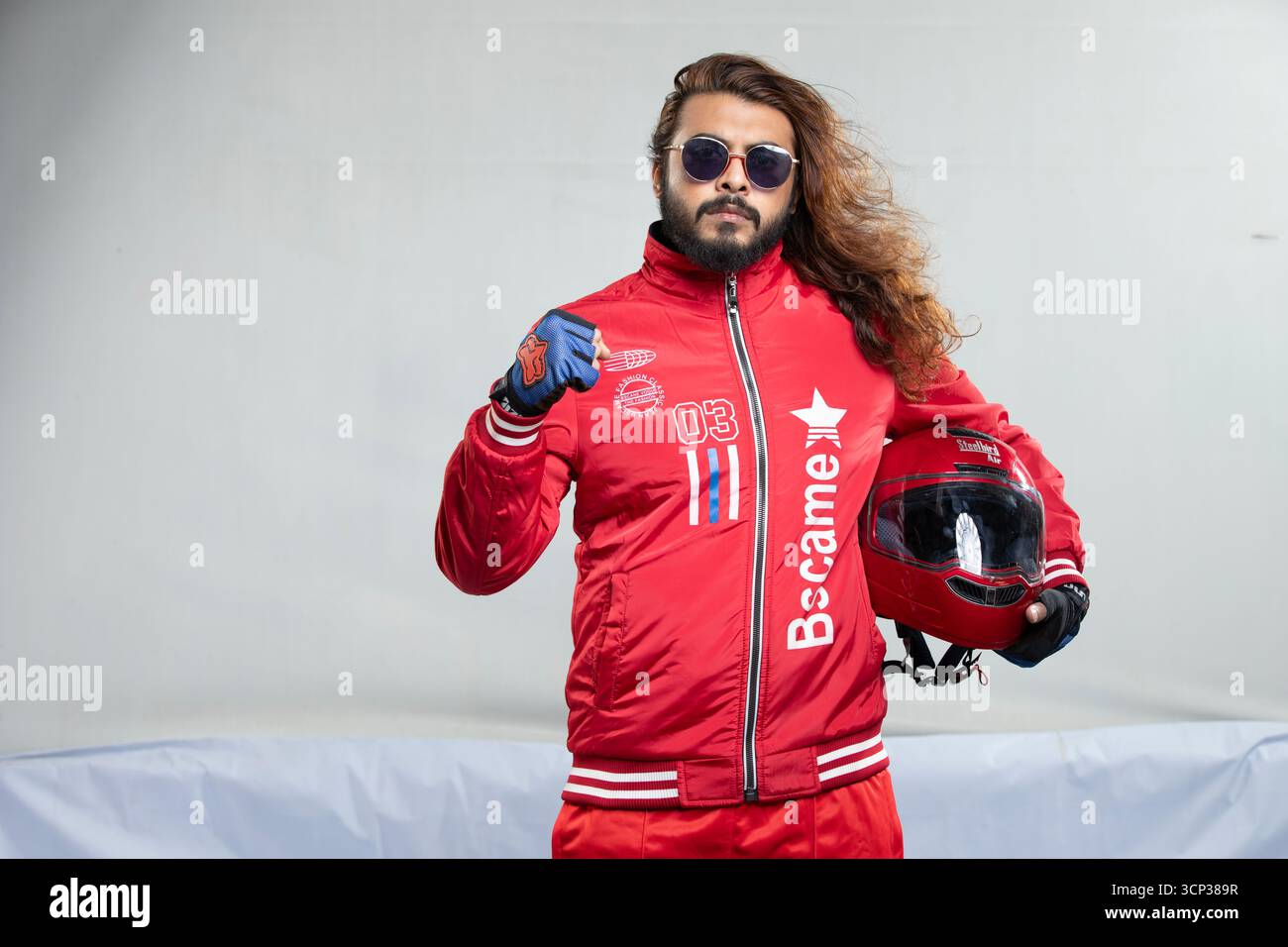 Uomo sicuro di sé con capelli lunghi e occhiali da sole in posa con un casco da moto rosso Foto Stock