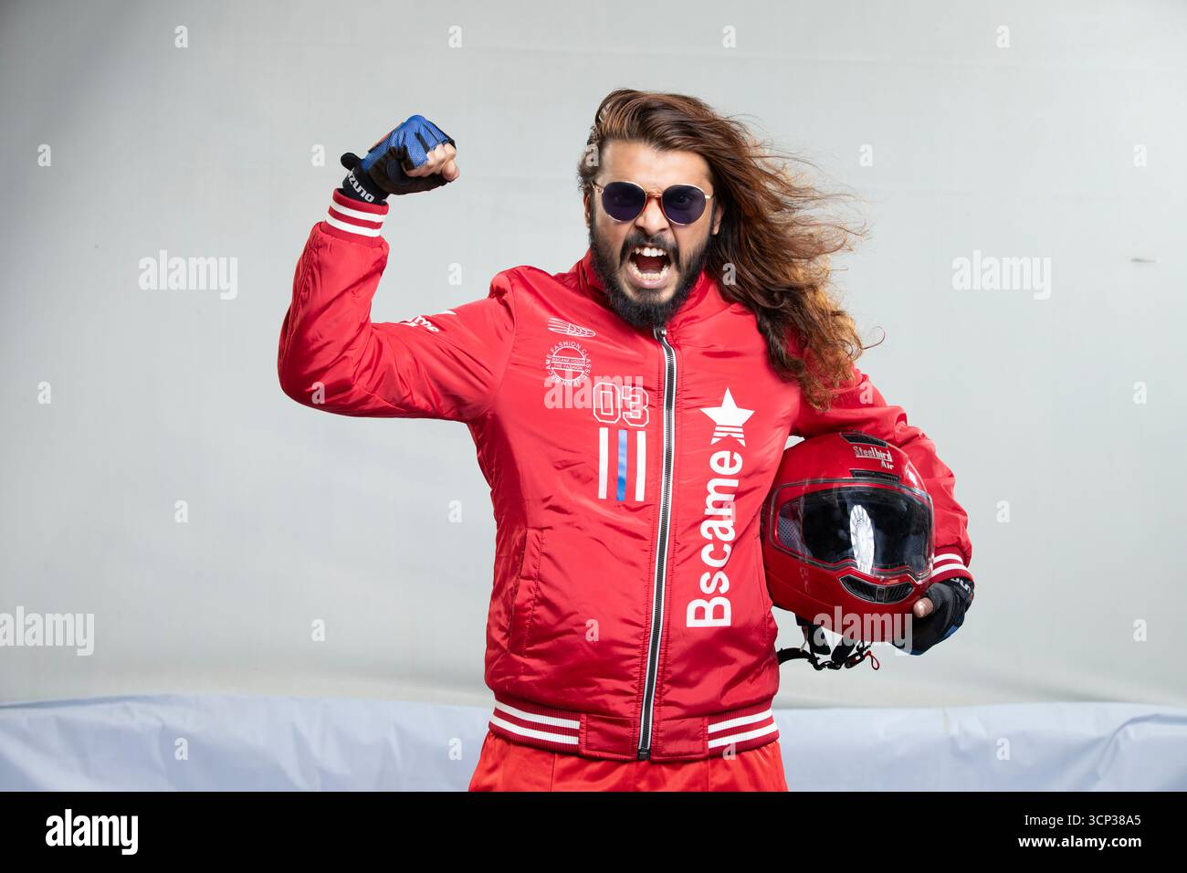 Uomo con capelli lunghi e occhiali da sole in una giacca rossa in posa con casco da moto Foto Stock