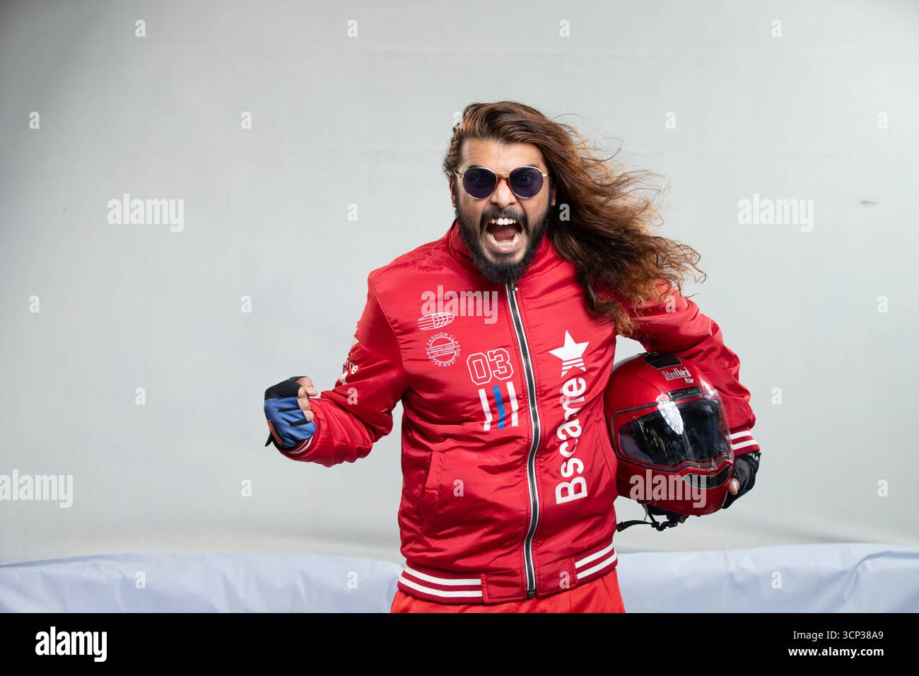 Uomo con capelli lunghi e occhiali da sole in una giacca rossa in posa con casco da moto Foto Stock
