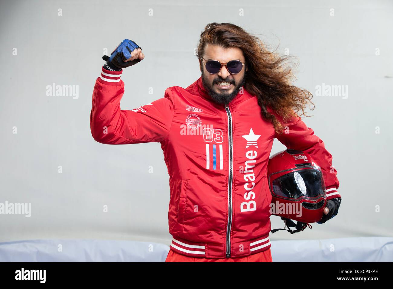 Uomo con capelli lunghi e occhiali da sole in una giacca rossa in posa con casco da moto Foto Stock