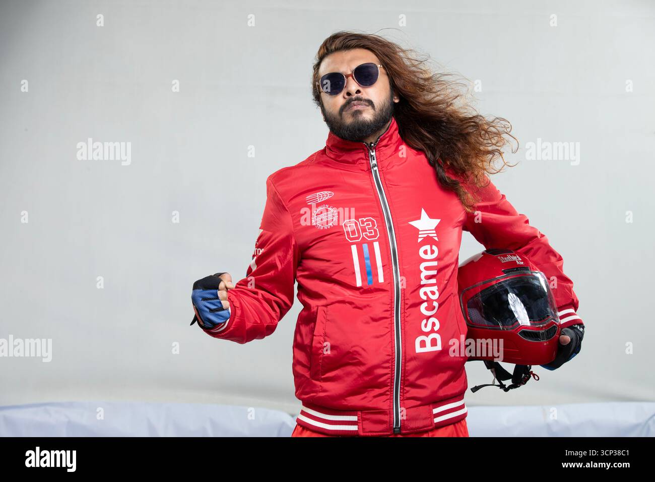 Uomo sicuro di sé con capelli lunghi e occhiali da sole in posa con un casco da moto rosso Foto Stock