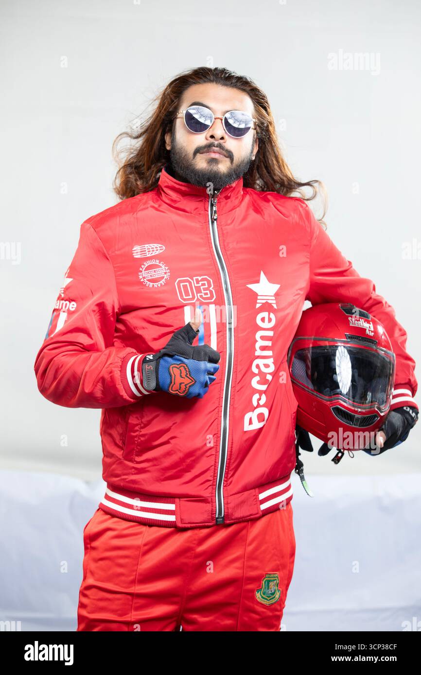 Uomo sicuro di sé con capelli lunghi e occhiali da sole in posa con un casco da moto rosso Foto Stock