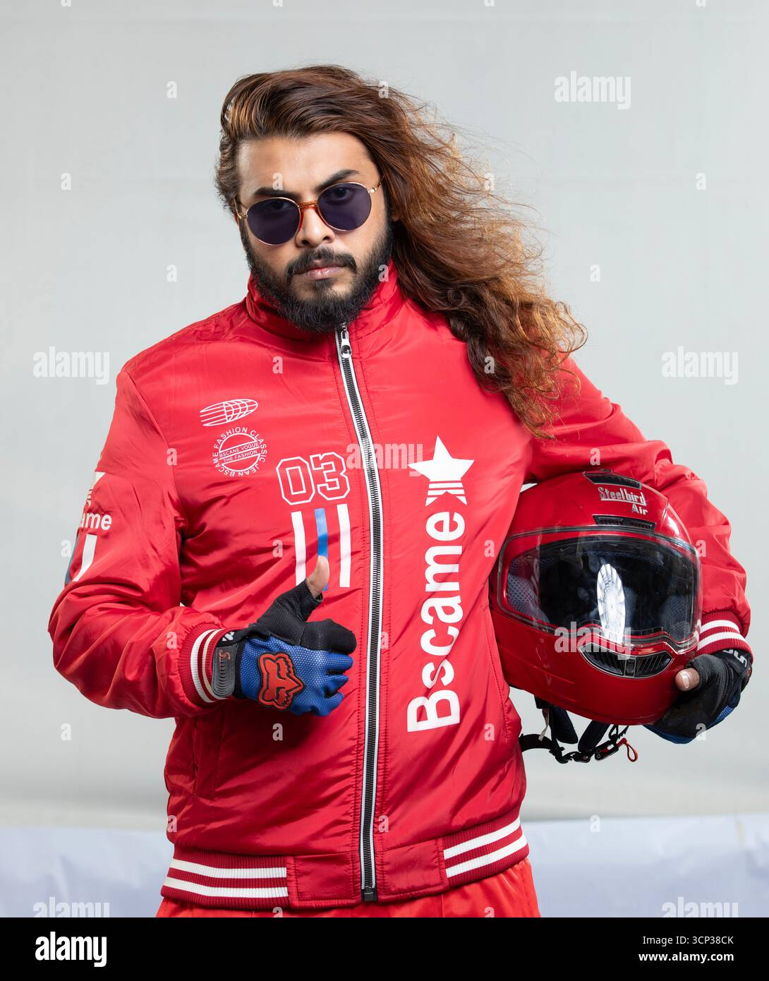 Uomo sicuro di sé con capelli lunghi e occhiali da sole in posa con un casco da moto rosso Foto Stock