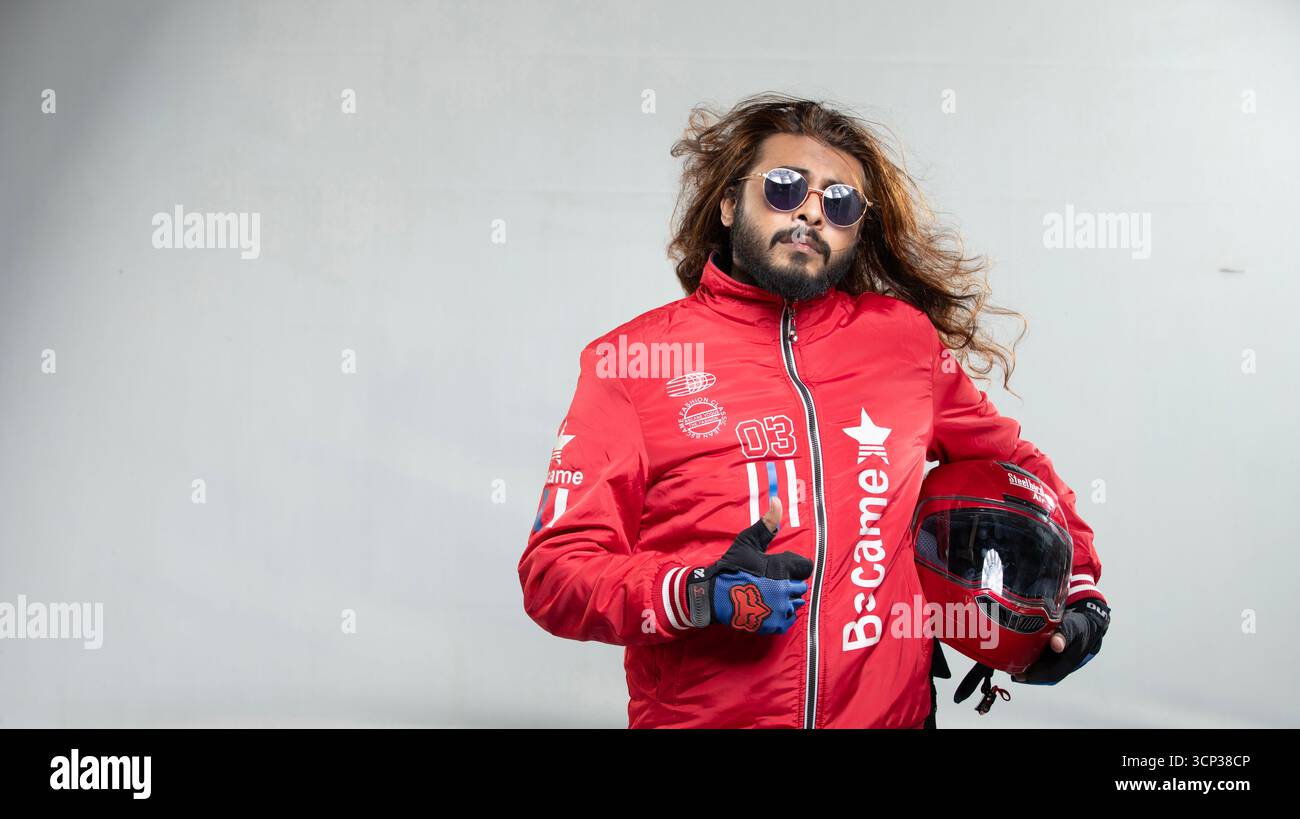 Uomo sicuro di sé con capelli lunghi e occhiali da sole in posa con un casco da moto rosso Foto Stock