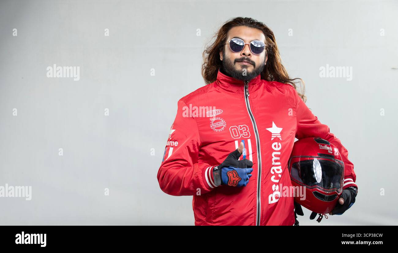 Uomo sicuro di sé con capelli lunghi e occhiali da sole in posa con un casco da moto rosso Foto Stock