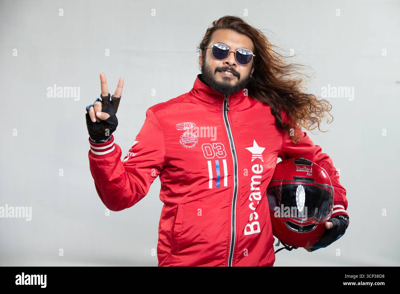 Uomo con capelli lunghi e occhiali da sole che dà un segno di pace mentre tiene in mano un casco da moto rosso Foto Stock