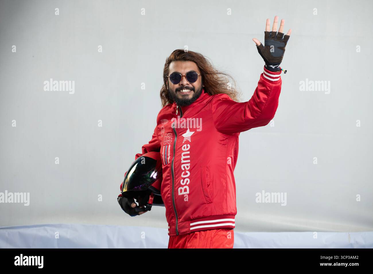 Uomo allegro con capelli lunghi e occhiali da sole che mostra Hi con un'onda amichevole tenendo in mano un casco nero Foto Stock