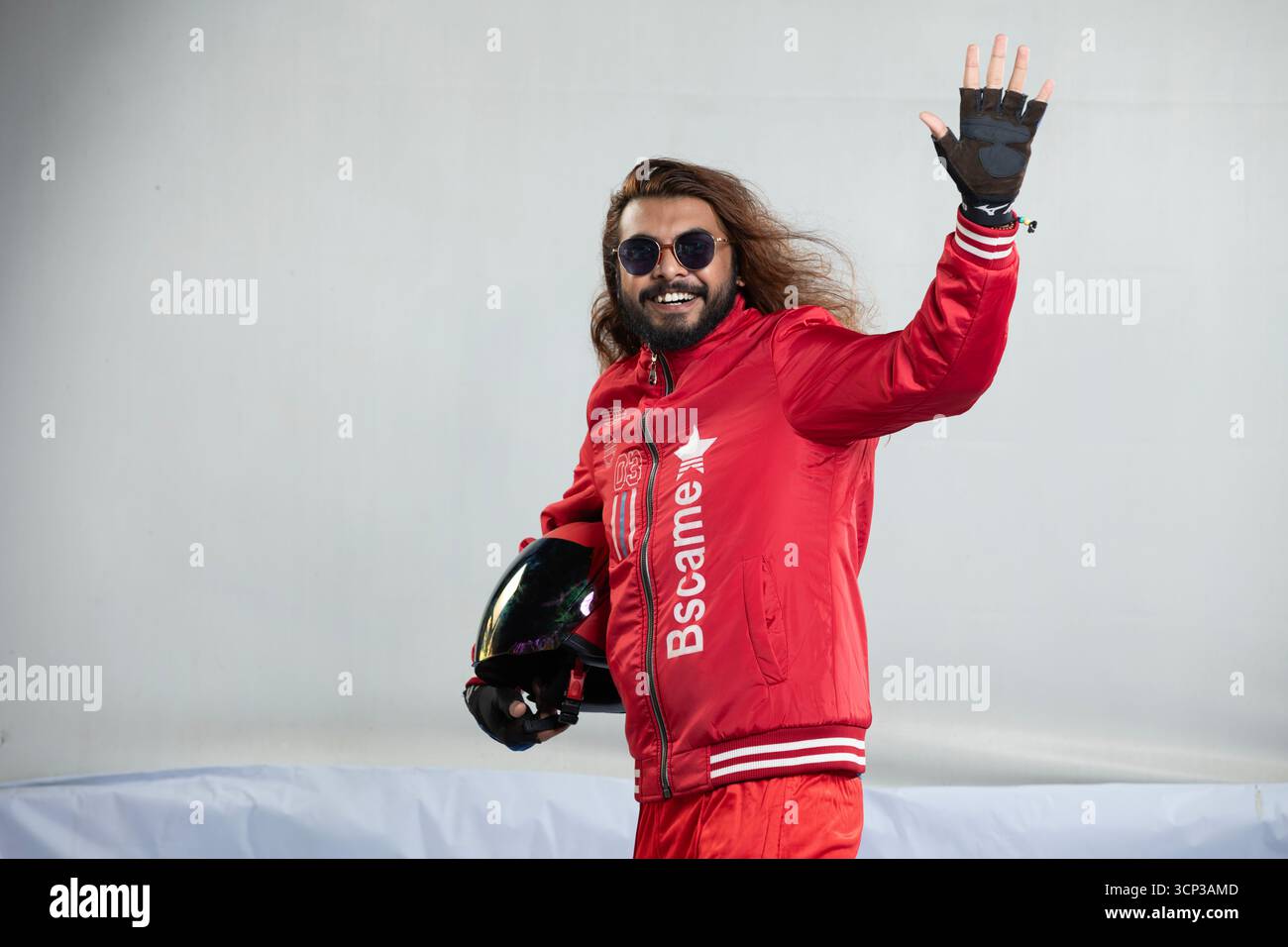 Uomo allegro con capelli lunghi e occhiali da sole che mostra Hi con un'onda amichevole tenendo in mano un casco nero Foto Stock