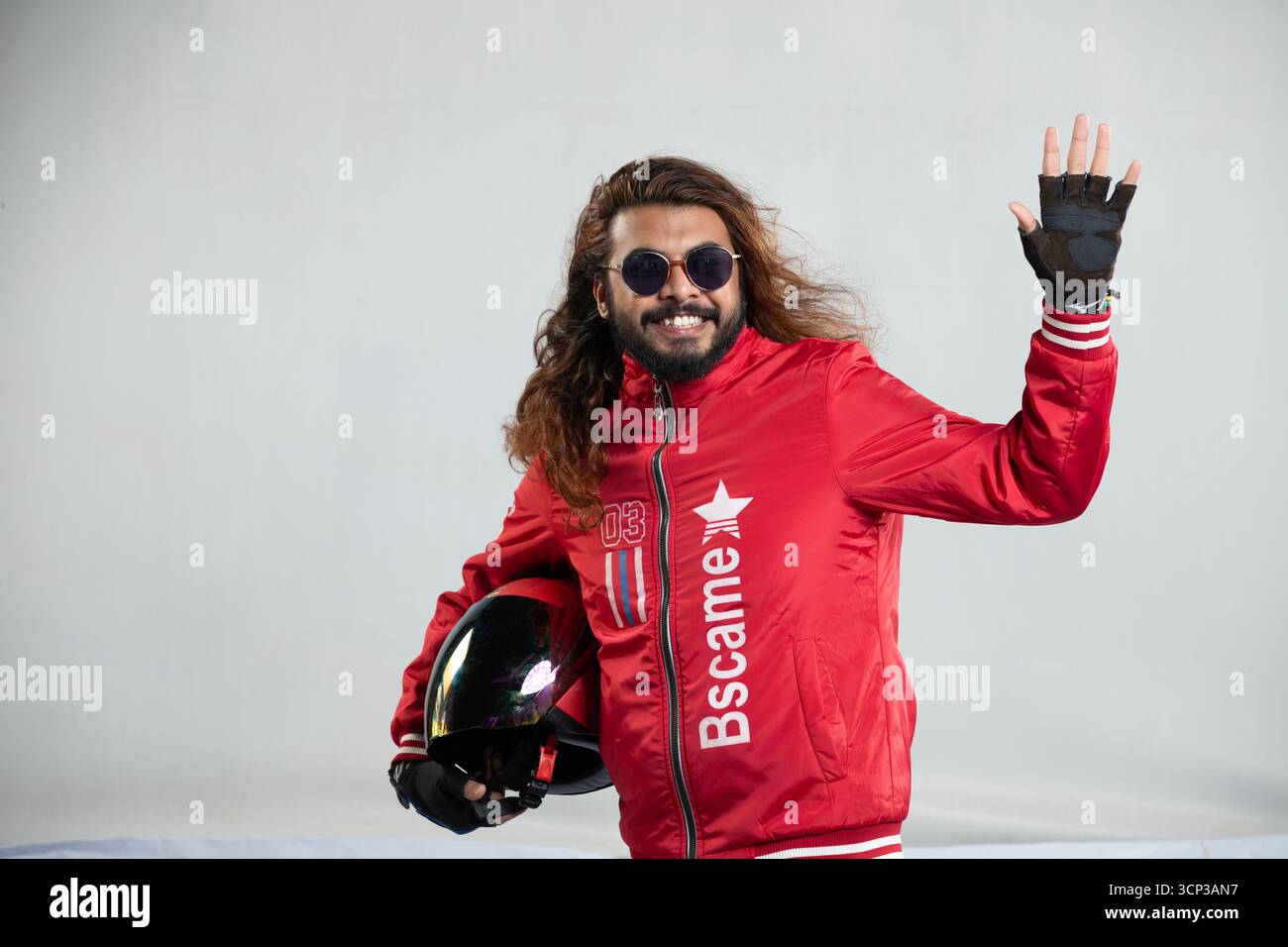 Uomo allegro con capelli lunghi e occhiali da sole che mostra Hi con un'onda amichevole tenendo in mano un casco nero Foto Stock