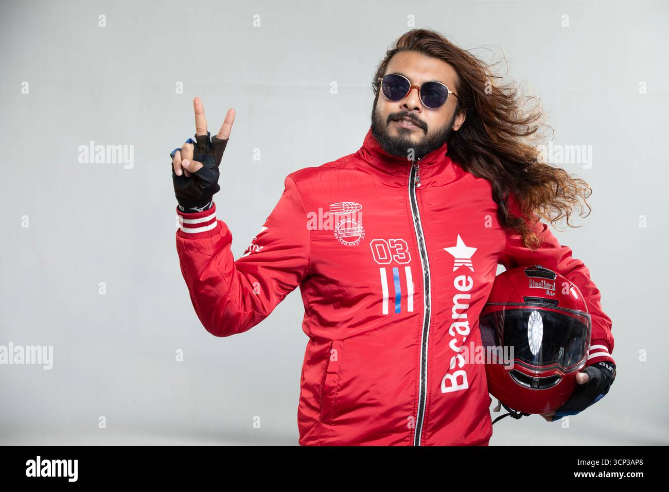 Confident Motor Car Racer Champion con capelli lunghi e occhiali da sole in una giacca rossa audace che celebra la vittoria con un gesto V-Sign Foto Stock