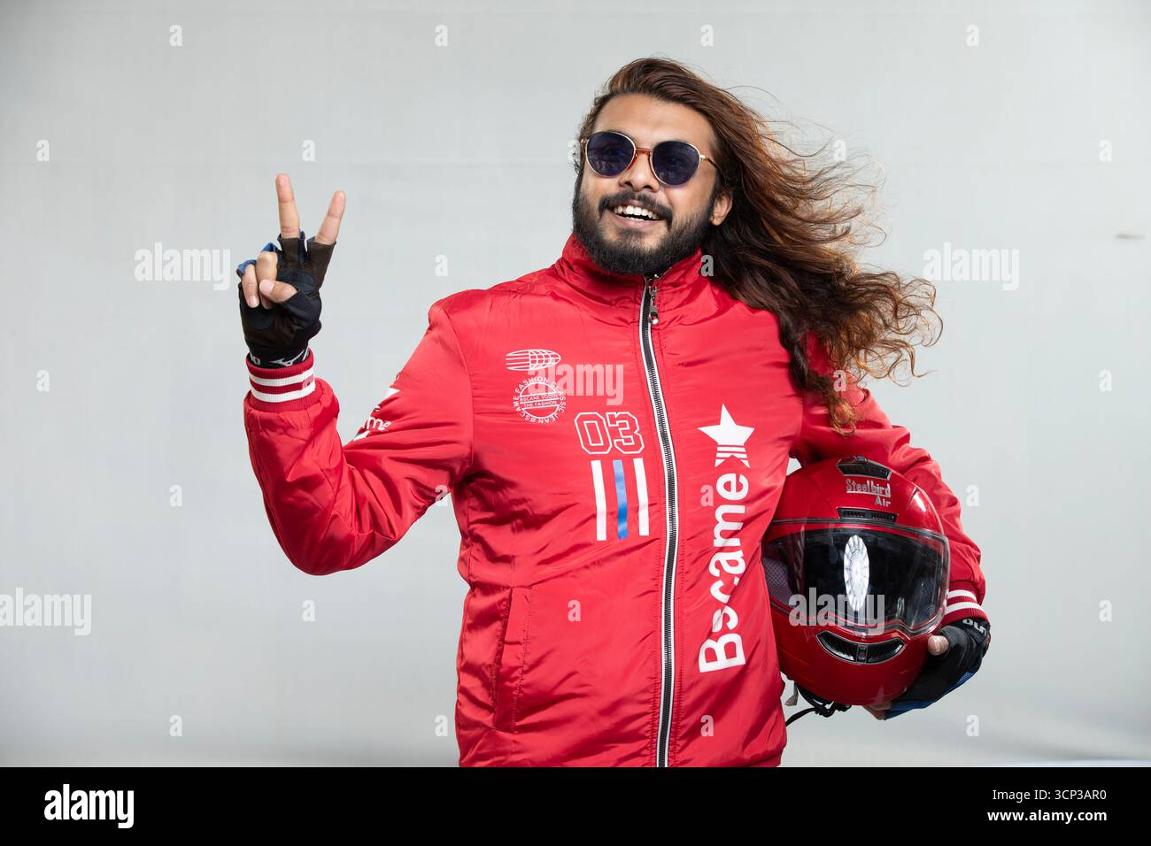 Confident Motor Car Racer Champion con capelli lunghi e occhiali da sole in una giacca rossa audace che celebra la vittoria con un gesto V-Sign Foto Stock