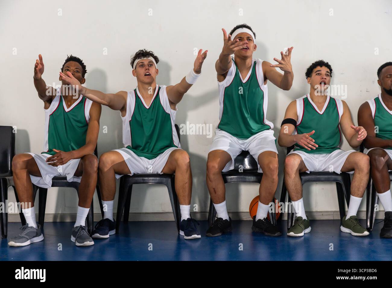 Compagni di squadra maschi in uniforme da basket urlano mentre si siedono sulle sedie a campo con il pallacanestro Foto Stock