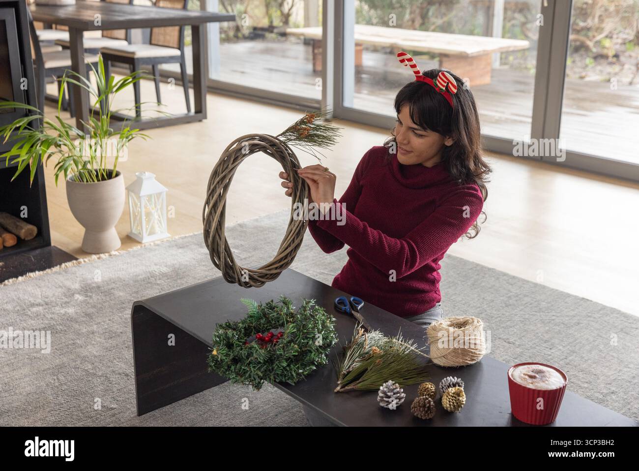 Donna che indossa un maglione seduto sul pavimento di casa, assemblando una corona con base vite, spago, spazio copia Foto Stock