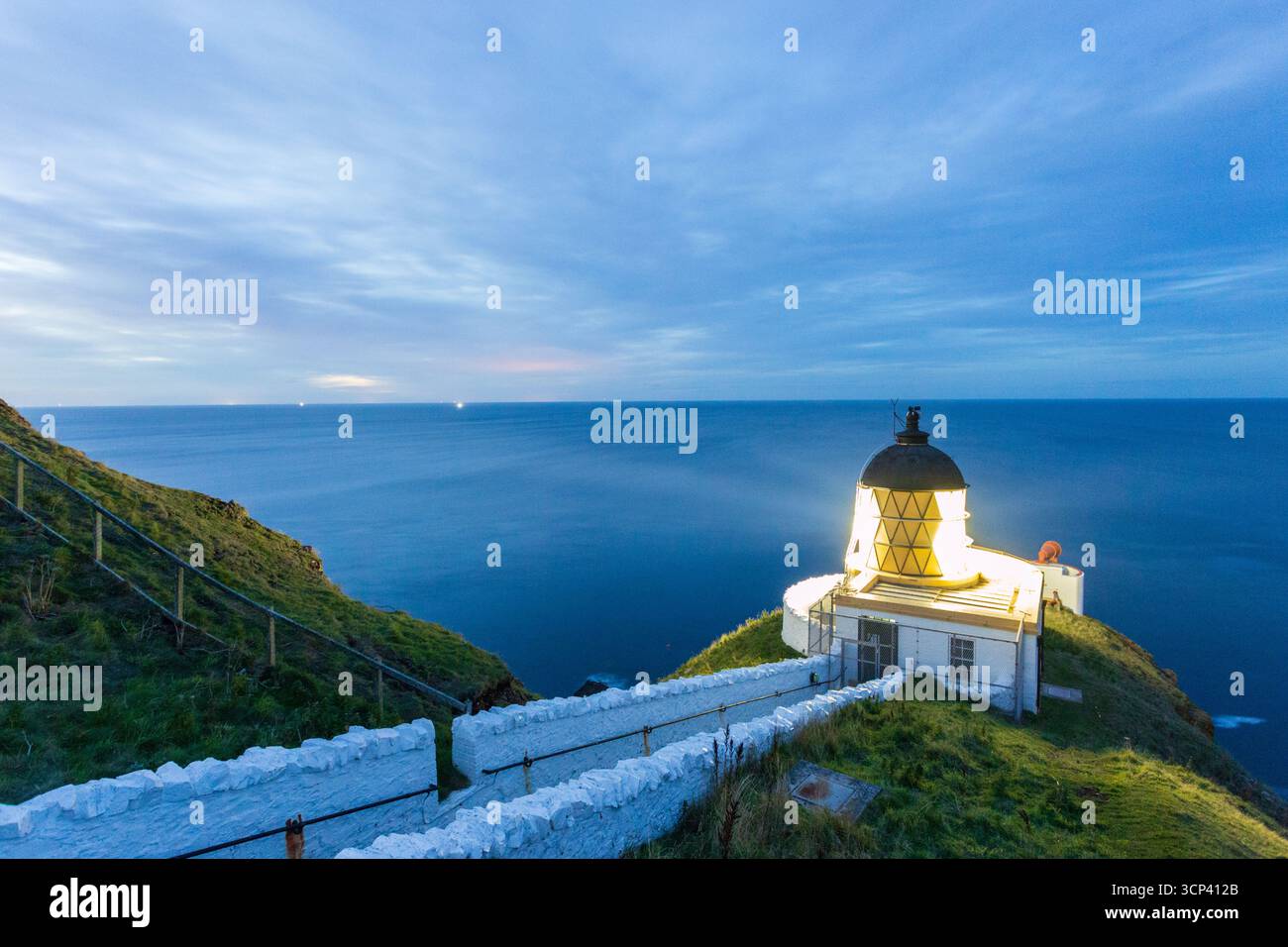 Vista di un faro radioso arroccato sulla cima di aspre scogliere, la sua luce che taglia attraverso il crepuscolo sul tranquillo mare blu scuro, St Abbs Head, Scozia. Foto Stock
