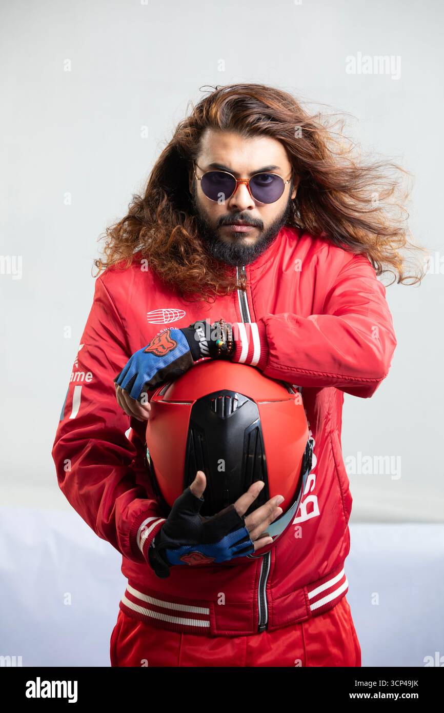 Un uomo con i capelli lunghi, la barba e gli occhiali da sole, che indossa una giacca rossa e tiene in mano un casco da moto e guanti. Foto Stock