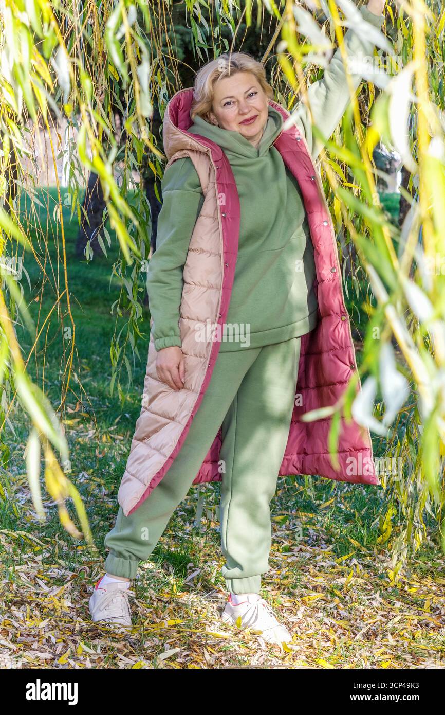 Una donna di mezza età con una tuta verde e un gilet beige e rosa sorridente si erge sulla strada tra i rami appesi di un salice. Brillante Foto Stock