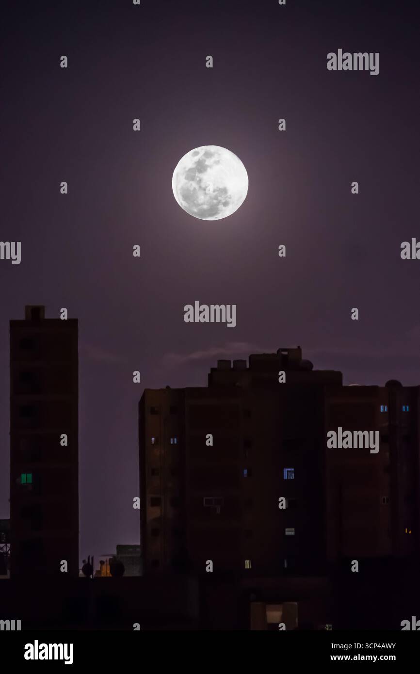 La luna piena splende luminosa nel cielo notturno sopra gli edifici della città, lo skyline urbano sotto la luce della luna in una serata limpida. Foto Stock