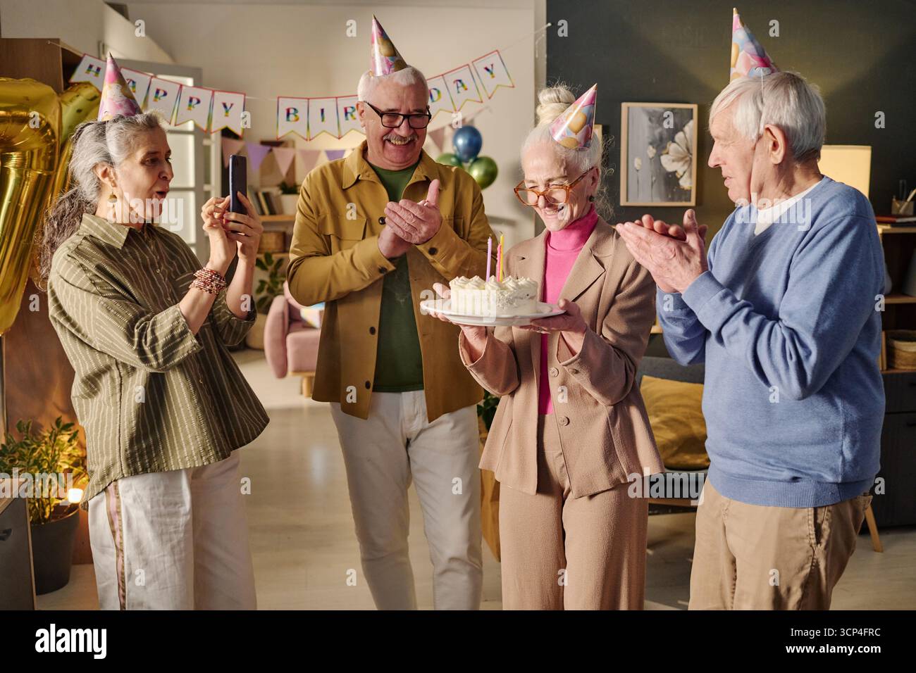 Donna anziana che tiene la torta di compleanno mentre gli amici applaudono sorridendo Foto Stock