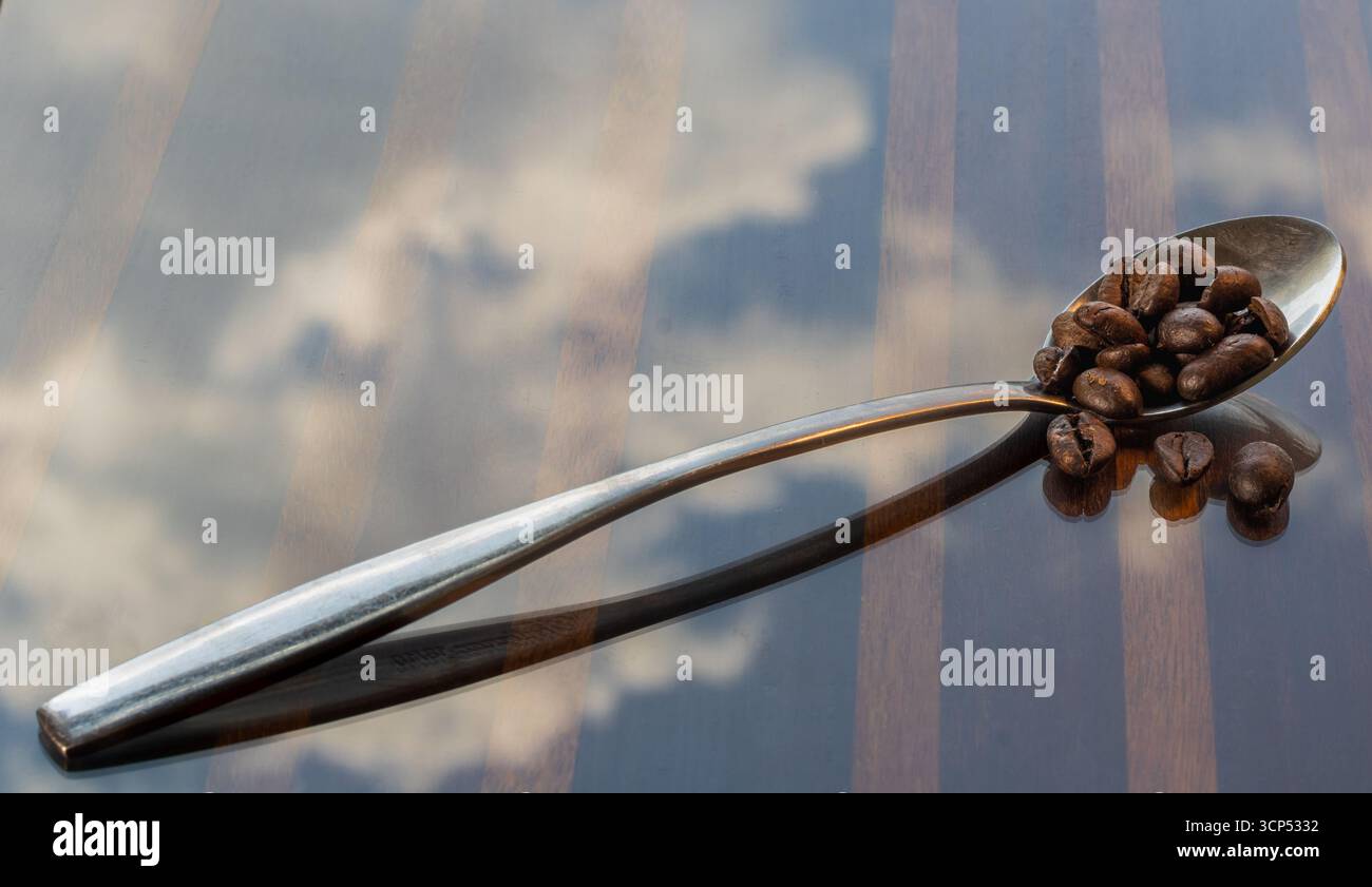 cucchiaio con chicchi di caffè sulla superficie riflettente, chicchi di caffè e riflessi del cielo Foto Stock