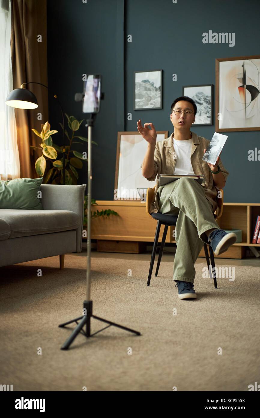 Giovane uomo asiatico seduto su una sedia tenendo un libro e gesti, mentre parla con lo smartphone su un treppiede registrando video nel moderno soggiorno con opere d'arte incorniciate sulla parete Foto Stock