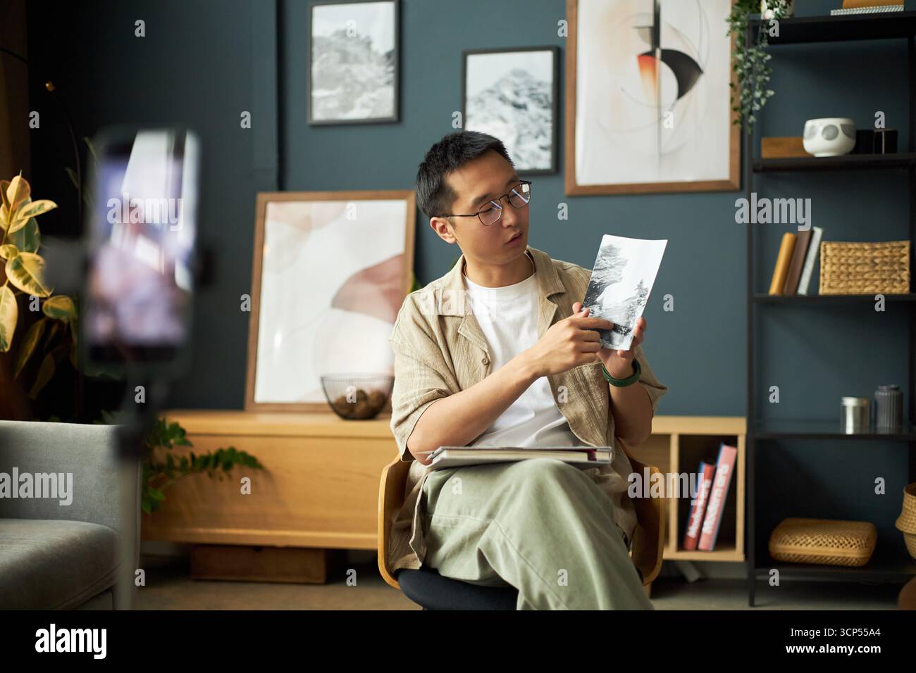 Giovane uomo asiatico seduto e che spiega la fotografia mentre si muove verso l'immagine stampata, tiene il notebook in grembo, lo smartphone su treppiede registra video in una stanza moderna Foto Stock