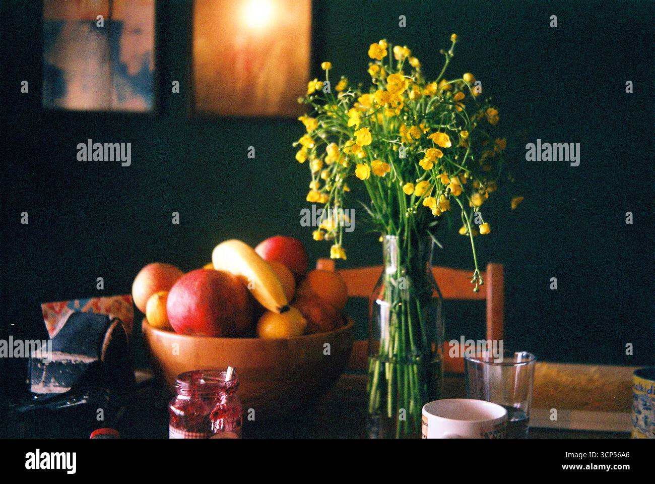 Natura morta analogica di fiori gialli e una ciotola di frutta contro una parete verde, illuminata dalla luce naturale. Ambiente domestico semplice e calmo. Foto Stock