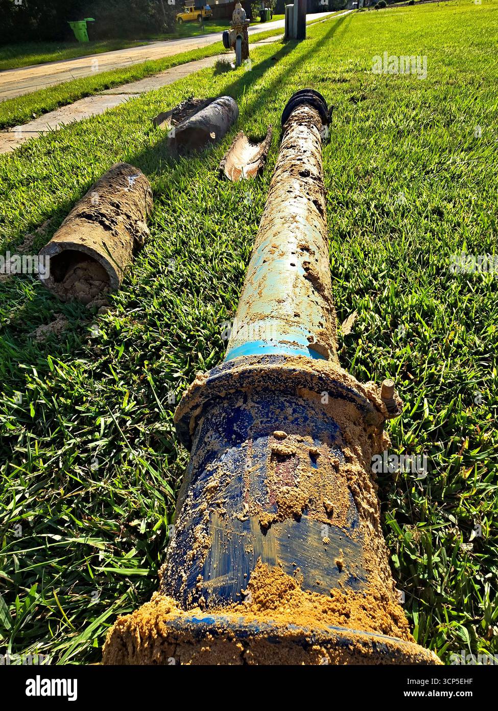 Un colpo ravvicinato di un tubo d'acqua rotto coperto di fango, poggiato su un letto di erba verde, con una strada sullo sfondo. Foto Stock