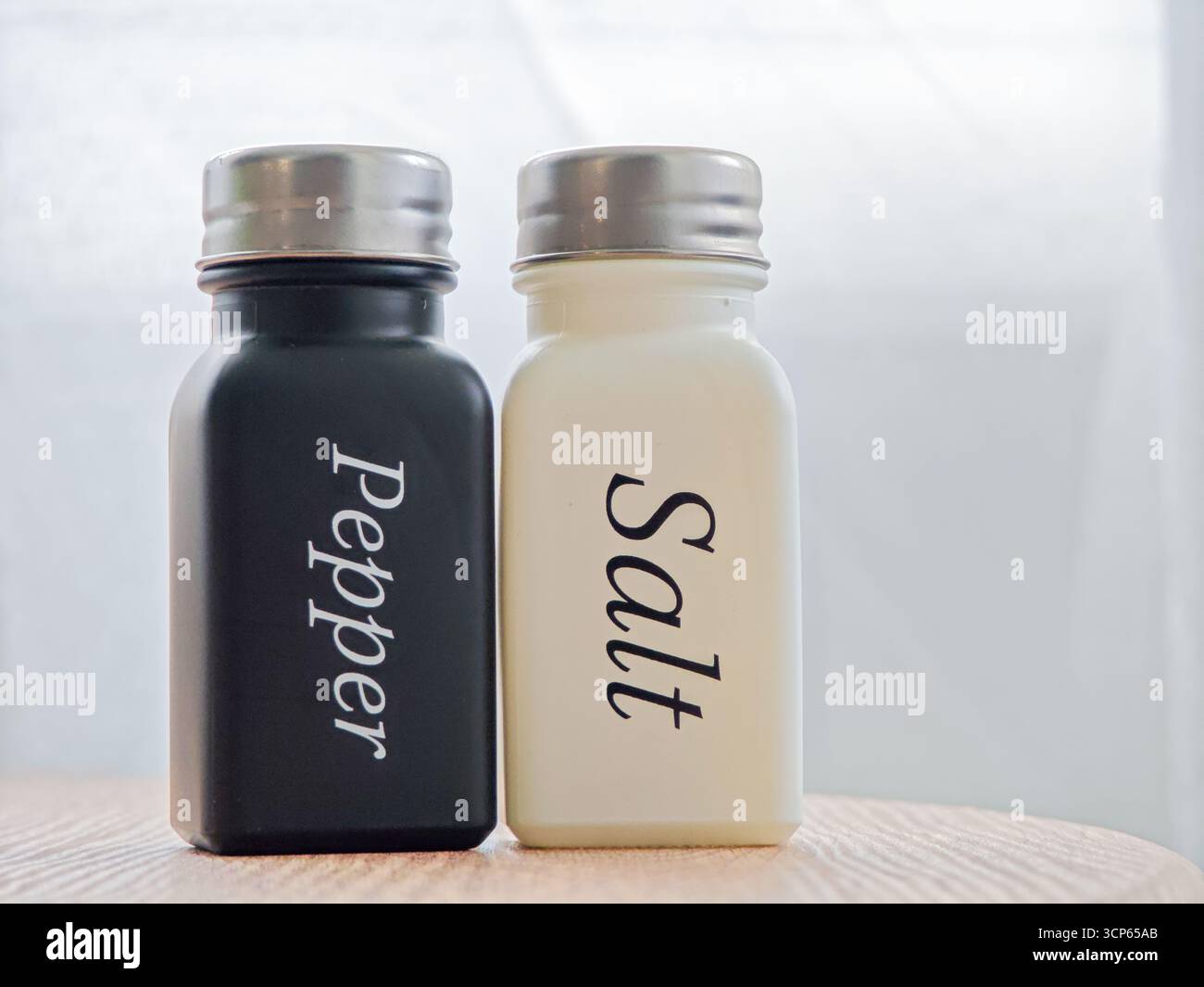 Su un tavolo di legno, due contenitori di condimenti si trovano fianco a fianco. Uno è nero etichettato Pepper, mentre l'altro è bianco etichettato Salt. Filtri luce solare Foto Stock