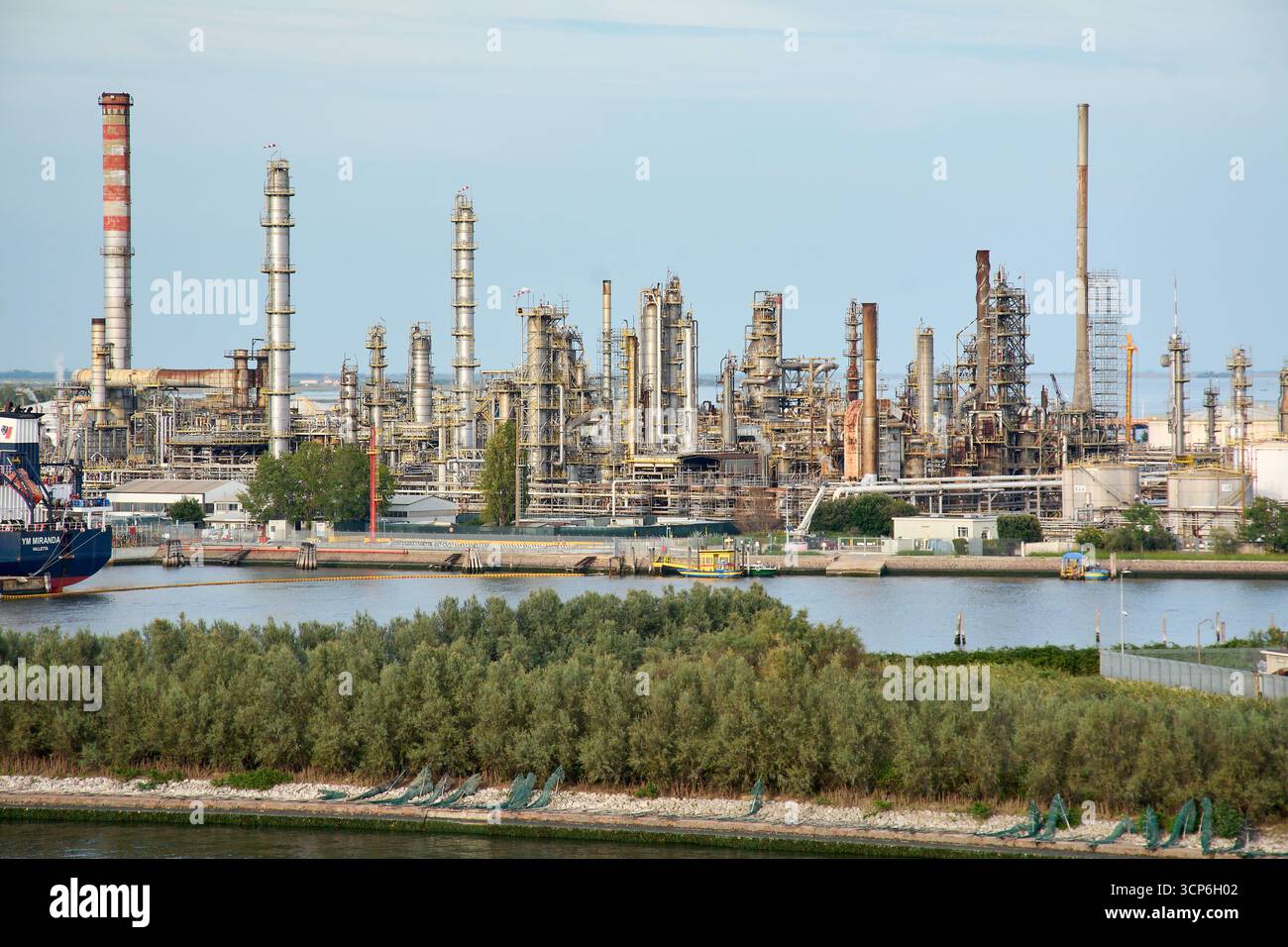 Venezia. Italia - 24 settembre 2025: Un'estesa raffineria di petrolio e un impianto petrolchimico con numerose torri, tubi e fumogeni domina l'acqua Foto Stock