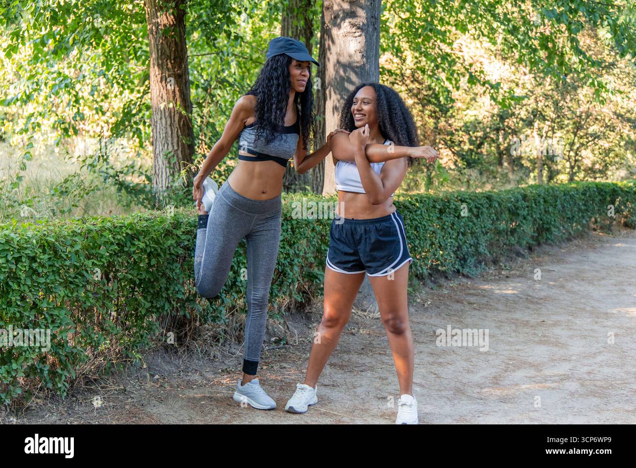 Due donne latine sorridenti che si allungano le gambe e le braccia prima di correre in un parco durante una giornata estiva di sole, promuovendo uno stile di vita sano e il benessere Foto Stock