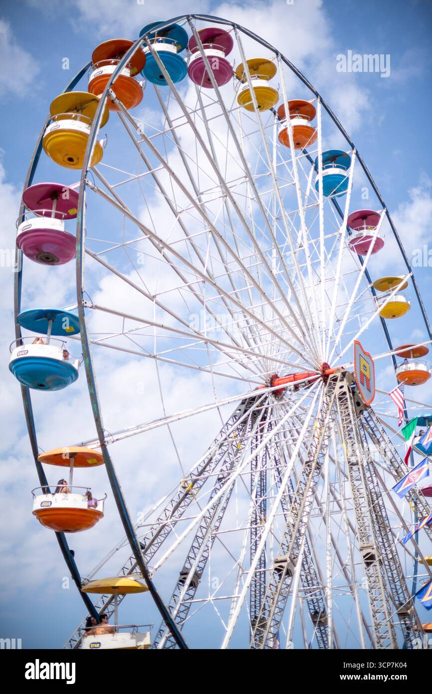 Ruota panoramica a Frederick Fairgrounds Frederick Maryland // FREDERICK, Maryland - Una ruota panoramica è raffigurata contro un cielo blu con nuvole sparse presso la Frederick Fairgrounds a Frederick, Maryland. Il giro di divertimento presenta una struttura in metallo bianco con numerose gondole colorate, tra cui giallo, arancione, rosa e blu, attaccata alla sua circonferenza. Alcune gondole sono etichettate con nomi come "CASEY", "JORDAN", "CAMERON", "JESSIE" e "RYAN". La ruota panoramica è un'attrazione importante, spesso associata a fiere e carnevali, che offre vedute panoramiche dell'area circostante. La F Foto Stock
