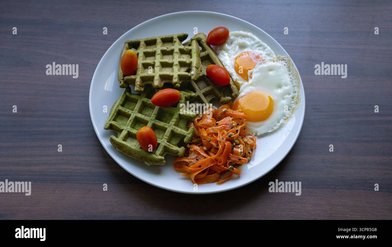 Piatto della colazione con uova fritte, waffle verdi, pomodori ciliegini e insalata di carote, che illustrano uno stile di vita sano ed equilibrato. Foto Stock