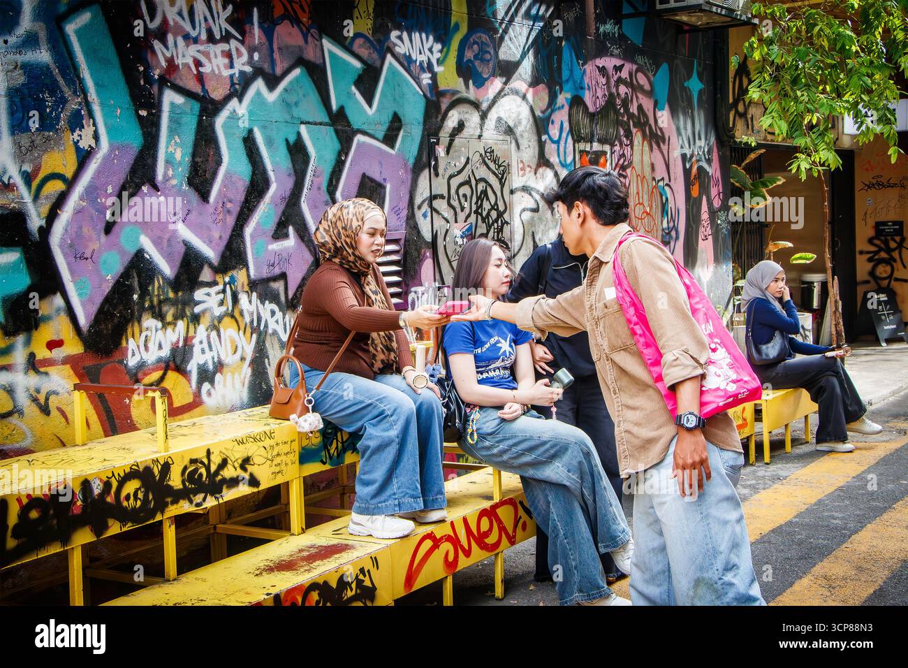 Un gruppo di giovani asiatici si siede e interagisce di fronte a un muro coperto di graffiti su una strada della città, arte e stile di vita urbano di kuala lumpur, malesia Foto Stock