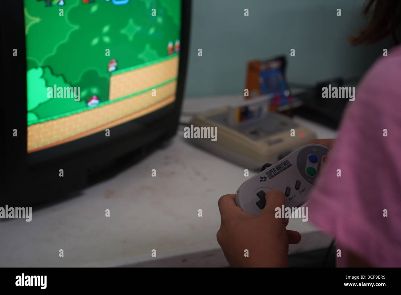 Un'immagine nostalgica di un Super Nintendo (SNES) vintage e di un controller su una scrivania. Una ragazza gioca un classico su un vecchio TV CRT, catturando i videogame degli anni '90. Foto Stock