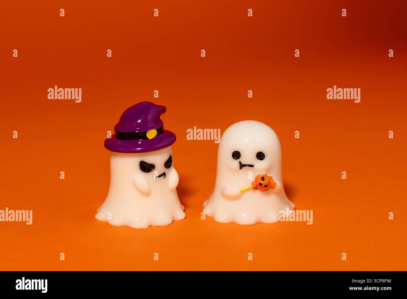 Un fantasma indossa un cappello da strega viola e sembra arrabbiato con il secondo fantasma, mentre tiene in mano le caramelle di zucca, un aspetto tranquillo su un vivace sfondo arancione Foto Stock