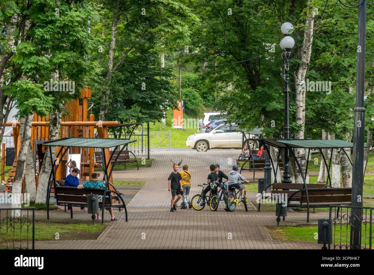 Bambini che camminano e cavalcano in bicicletta in un parco alberato, nella città di Korsakov, nell'isola di Sakhalin, Russia, con panchine ombreggiate, parco giochi in legno Foto Stock