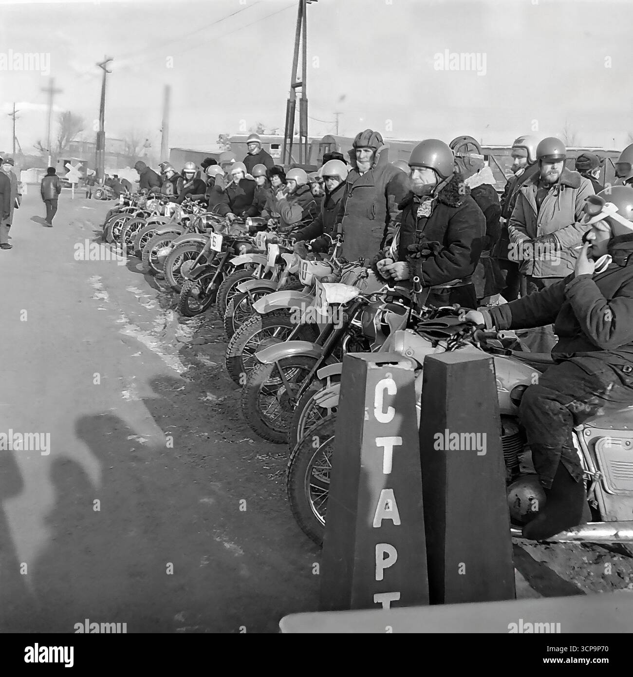 Una lunga fila di motociclisti sovietici si siede sulle loro moto, aspettando con tregua attesa l'inizio di una gara invernale di motocross a Sloviansk, RSS Ucraina, nel 1975. I piloti, vestiti con pesanti outfit invernali, sono allineati dietro i coni "START" sulla pista ghiacciata, a dimostrazione della grande popolarità di questo evento motoristico di base. Questa suggestiva fotografia in bianco e nero cattura la messa a fuoco collettiva e i momenti di eccitazione prima dell'inizio della competizione Foto Stock
