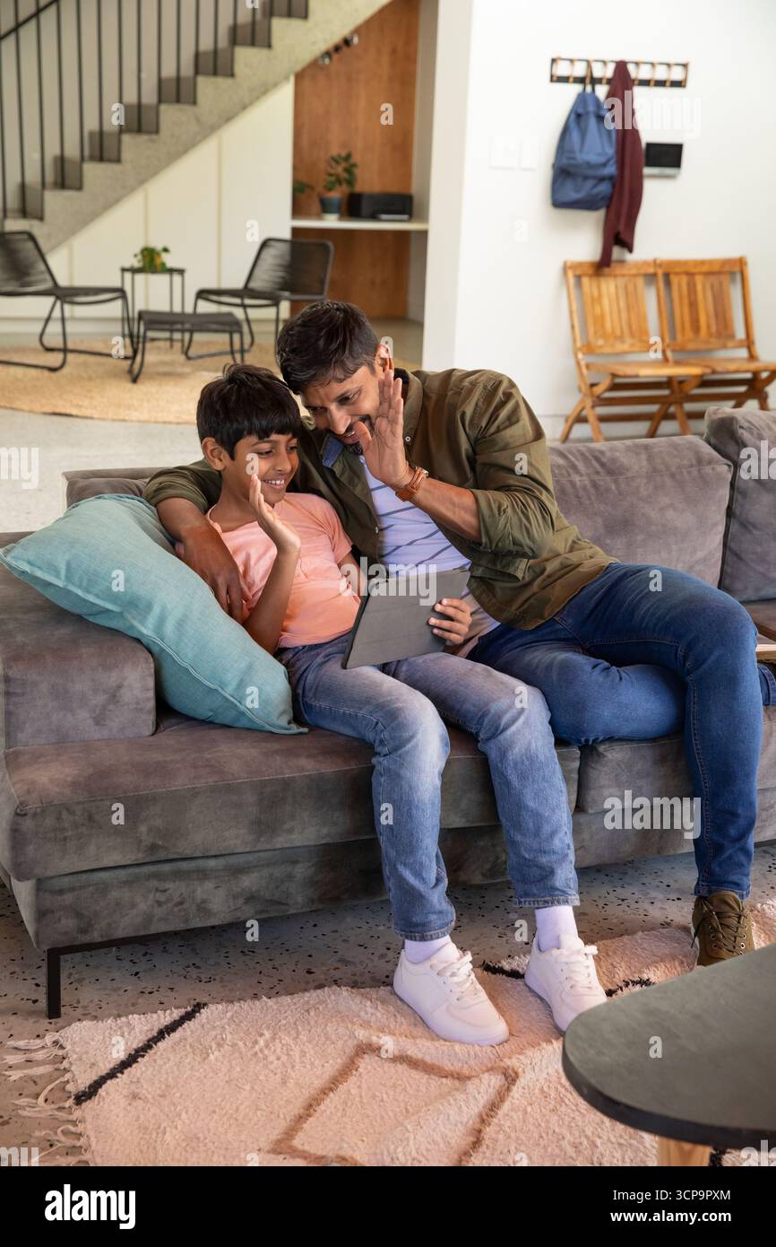 Padre e figlio dell'asia meridionale che sventolano al tablet mentre si siedono sul divano in soggiorno Foto Stock