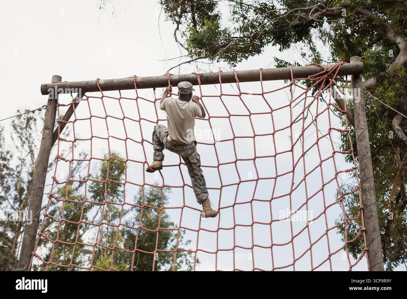 Soldato afroamericano maschio in camo arrampicata rete di corda rossa che raggiunge la trave di legno su percorso ad ostacoli Foto Stock