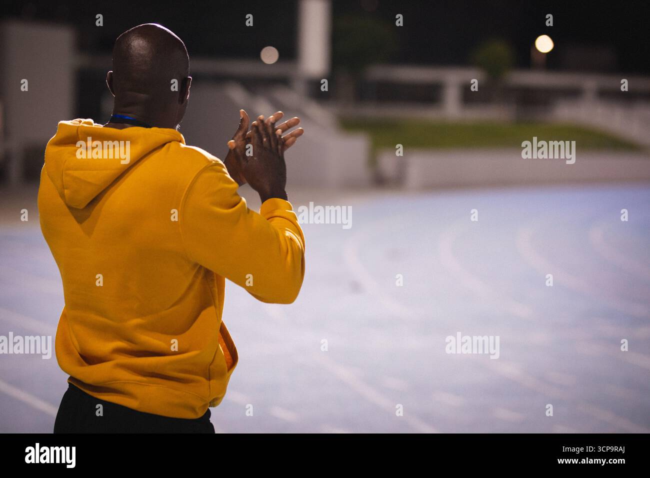 Uomo afroamericano con felpa gialla che batte sulle piste sotto le luci dello stadio, copia spazio Foto Stock
