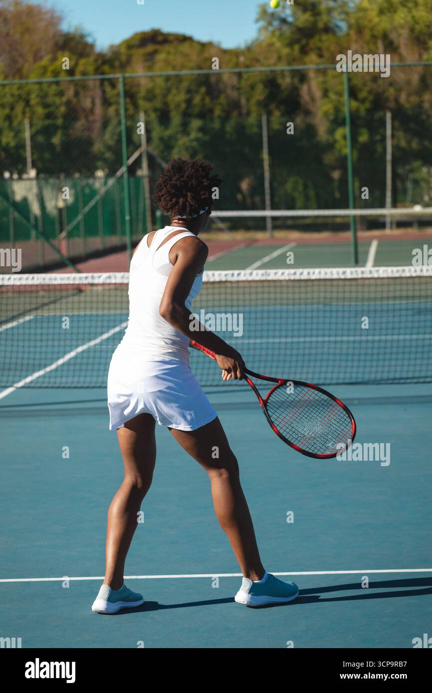 Donna afroamericana che indossa un abito da tennis con racchetta che si prepara a colpire la palla sul campo da tennis Foto Stock