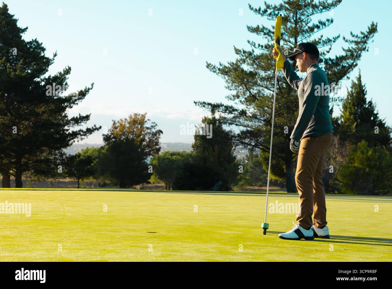 Golfista maschio con i guanti in piedi sul putting green tenendo il flagstick, focalizzandosi sulla palla in tazza, copia spazio Foto Stock