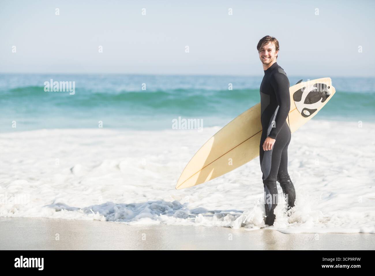Surfista maschio adulto che indossa una muta con tavola da surf in piedi sulla costa mentre le onde si infrangono, spazio per copiare Foto Stock
