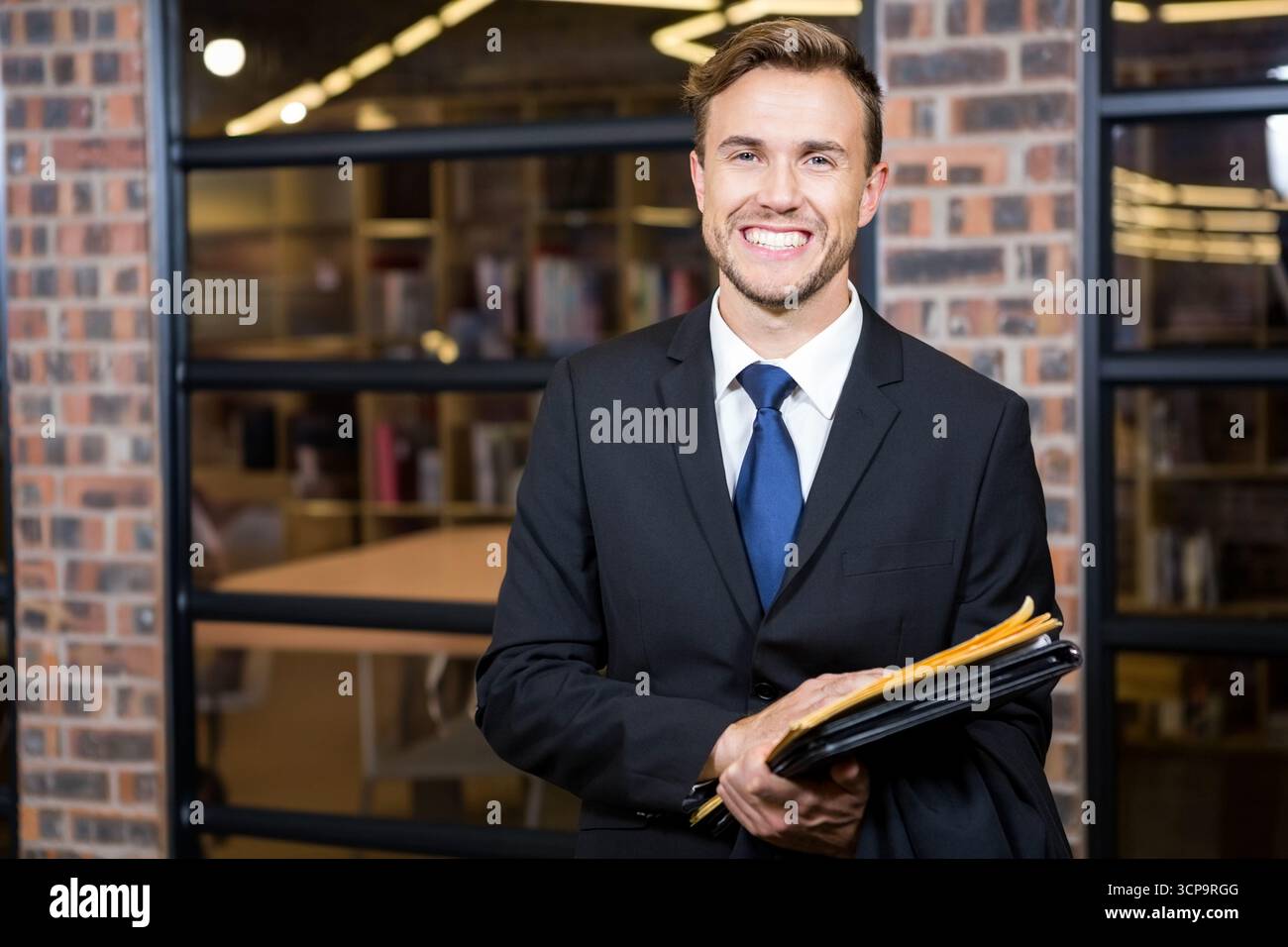 Uomo d'affari che indossa una tuta nera e una cravatta blu che tiene le cartelle vicino alla partizione di vetro in un ufficio moderno Foto Stock