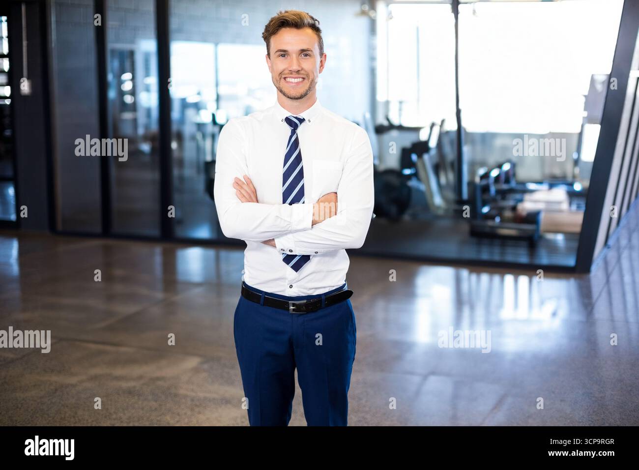 Uomo d'affari in piedi con le braccia incrociate in palestra con pareti divisorie in vetro e cyclette Foto Stock