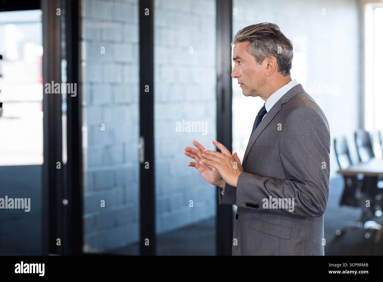 Uomo d'affari in piedi e che parla con pareti divisorie in vetro vicino al tavolo da conferenza e alle sedie in ufficio Foto Stock