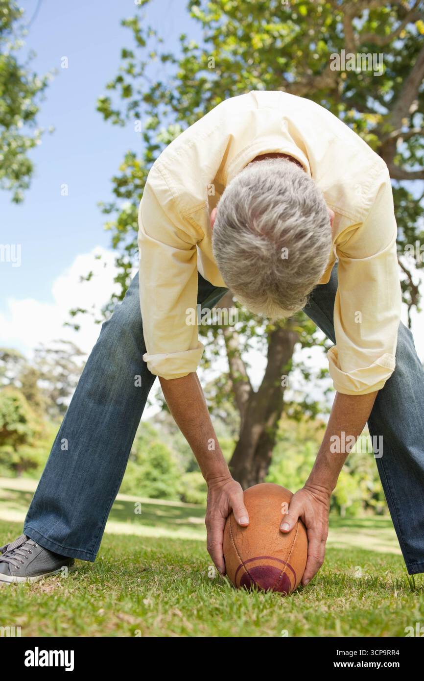 Maschio che si piega sopra il posizionamento del football americano sull'erba del parco sotto il cielo blu con una camicia gialla pallida Foto Stock