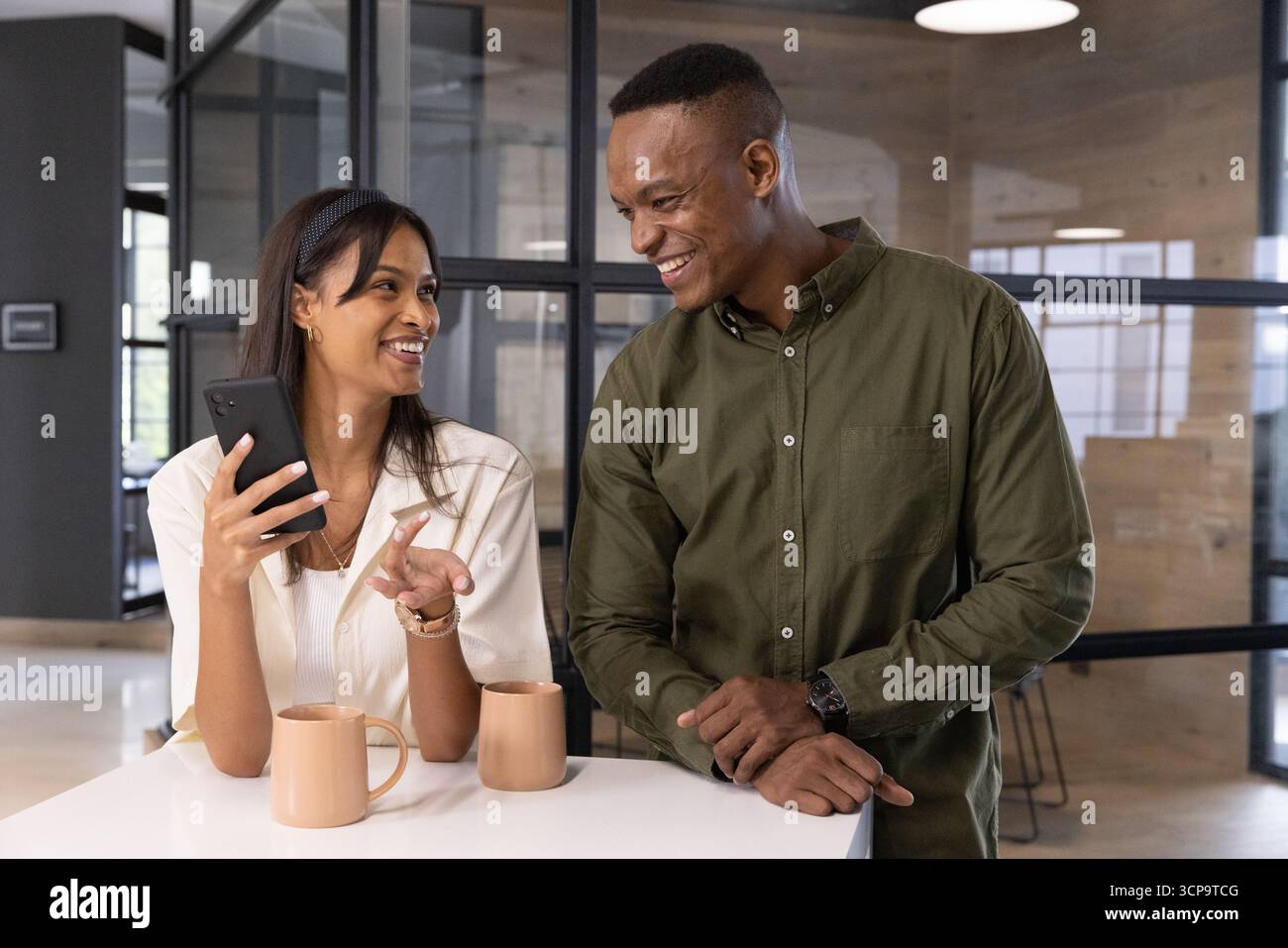 Diversi colleghi appoggiati al piano di lavoro nella sala colazione dell'ufficio che chiacchierano con lo smartphone, due tazze Foto Stock